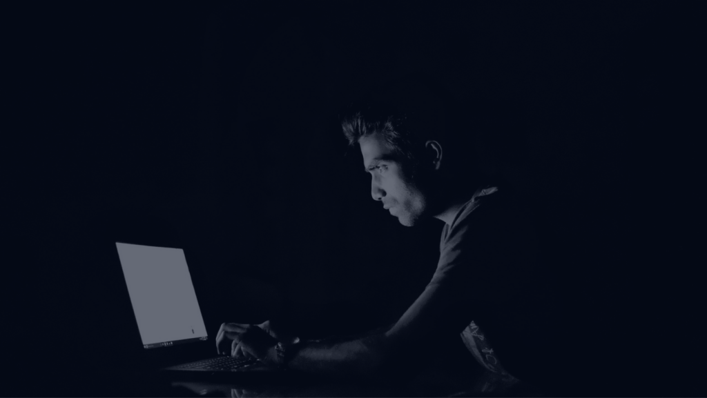 The picture shows a young man sitting at a laptop in a dark room spreading disinformation using ChatGPT. The image is in black and white with a really high contrast. 